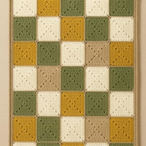 Puede incluir: Una manta de ganchillo hecha a mano con un diseño de tablero de ajedrez. Los cuadrados alternan crema, verde oliva, amarillo mostaza y beige. La manta tiene un borde beige.