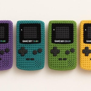 Puede incluir: Cuatro consolas de juegos portátiles Game Boy Color de crochet en morado, verde azulado, verde y amarillo.