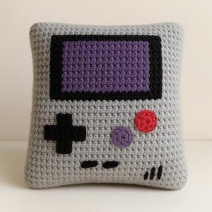 May include: A crocheted pillow in the shape of a retro handheld game console. The pillow features a gray body with a purple screen, black border, and black directional pad. It also has red and purple buttons, and black indicator lines.