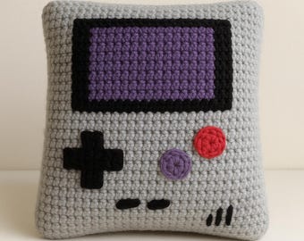 May include: A crocheted pillow in the shape of a retro handheld game console. The pillow features a gray body with a purple screen, black border, and black directional pad. It also has red and purple buttons, and black indicator lines.