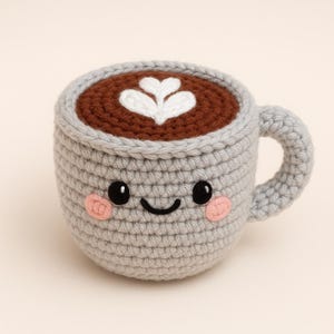 May include: A grey crocheted coffee cup with a brown crocheted coffee and a white crocheted foam design. The cup has a happy face with black eyes and a black smile. The cup has pink cheeks.