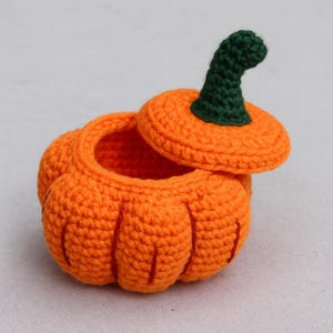 Puede incluir: Un recipiente con forma de calabaza de ganchillo naranja con tapa. La tapa está ligeramente entreabierta, revelando el interior hueco. El tallo es verde. El recipiente es probablemente hecho a mano y adecuado para objetos pequeños o como pieza decorativa.