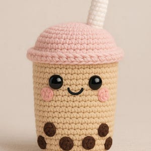 Puede incluir: Una taza de té boba de ganchillo con una cara sonriente. La taza es de color beige con bobas marrones, una tapa rosa, ojos negros y mejillas rosas. Una pajita blanca se extiende desde la parte superior.