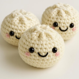 May include: Three crocheted stuffed dumplings with smiling faces. Each dumpling is white with black eyes and a black smile. The dumplings have pink cheeks.