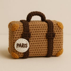 Può includere: Una valigia marrone all'uncinetto con cinghie, manico e angoli marrone scuro. Una toppa circolare bianca con la scritta "PARIS" è sul davanti. La valigia è un oggetto decorativo.