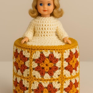 May include: A vintage doll with blonde hair and blue eyes wearing a cream crocheted outfit and hat. The doll sits on a crocheted toilet roll cover with a floral pattern in cream, orange, and gold.