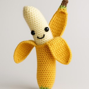 May include: A handmade crochet banana with a smiling face. The banana is yellow with a cream-colored center and brown and green accents. The banana peel is open, revealing the smiling face with black eyes and a curved mouth.