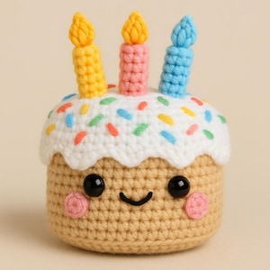 May include: A crocheted birthday cake plush toy with a smiling face, white icing, colourful sprinkles, and three candles in yellow, pink, and blue.