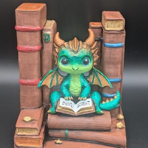 May include: A whimsical figurine of a green and teal dragon reading a book, nestled among faux brown books. The dragon has large, expressive eyes and copper-colored horns and wings. The base and surrounding books are painted in shades of brown and gold.
