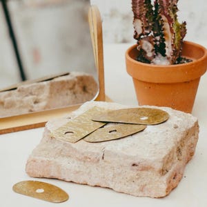 Op de afbeelding: Een stilleven met drie messing metalen stukken met een gat, rustend op een lichtroze steen. Een vierde stuk ligt apart. Een kleine cactus in een terracotta pot en een spiegel met houten frame staan op de achtergrond.