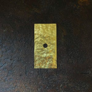 Puede incluir: Una placa rectangular de metal dorado con un pequeño agujero circular centrado. La placa tiene una superficie texturizada y está colocada sobre un fondo oscuro y moteado. La estética general es minimalista e industrial.
