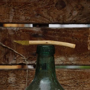 May include: A small, curved knife with a wooden handle and a brass blade, resting on top of a green glass bottle. The knife is positioned horizontally, with the blade facing left. The background is a wooden surface.