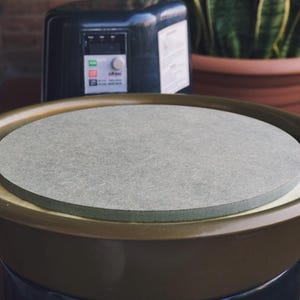 Op de afbeelding: Een pottenbakkerswiel met een grijs cirkelvormig werkoppervlak. Het wiel staat in een bronskleurig bassin. Een zwart bedieningspaneel en een plant in een pot staan op de achtergrond.