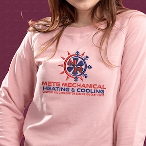 May include: A light pink sweatshirt with the text "METE MECHANICAL HEATING & COOLING" in red and blue. The design includes a logo with a fan and the letters "MM". The sweatshirt is a long-sleeved crew neck.