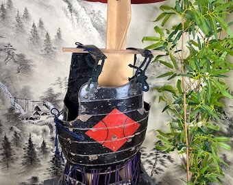 May include: A black lacquered samurai chest armor piece featuring a prominent red diamond emblem on the front, dark blue cord lacing, and a matching black conical hat with a red underside, all displayed on a wooden stand for structural support.