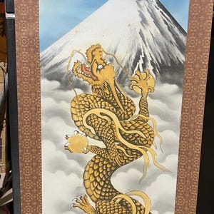 Puede incluir: Pintura en rollo vertical con un dragón dorado frente al monte Fuji. El dragón, con escamas detalladas, se alza sobre un cielo nublado y una montaña nevada. El rollo tiene bordes decorativos.