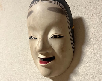 Könnte beinhalten: Eine traditionelle japanische Noh-Theatermaske mit einem heiteren Ausdruck. Die Maske hat ein blasses Gesicht, schwarzes Haar, dunkle Augenbrauen und rote Lippen. Die Maske ist an einer strukturierten weißen Wand befestigt.