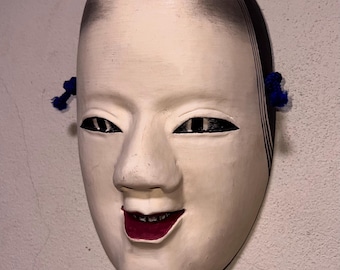 Könnte beinhalten: Eine traditionelle japanische Noh-Theatermaske mit einem lächelnden Ausdruck. Die Maske ist in Off-White bemalt, mit schwarzen Details für Augen, Haare und Mund. Blaue Quasten sind an den Seiten befestigt.