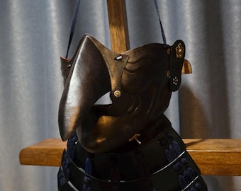 May include: A black Japanese samurai mask with a distinctive beak-like shape, displayed on a wooden stand. The mask features intricate details and is adorned with decorative elements. The armor is a traditional piece of Japanese history.