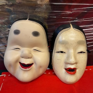 Peut inclure: Deux masques japonais traditionnels. Le masque de gauche est rond avec une expression souriante, des sourcils noirs et des lèvres rouges. Le masque de droite est ovale avec une expression sereine, des détails de cheveux noirs et des lèvres rouges.