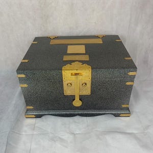 May include: A gray, rectangular box with gold-colored accents. The box features a gold clasp and decorative corner pieces. The box has a textured surface and is likely used for storage or display.
