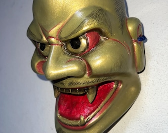 May include: A gold-coloured mask with a fierce expression. The mask features red accents around the eyes and mouth, with sharp teeth and a detailed design. The mask has a blue string attached to the ear.