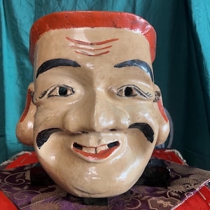 Peut inclure: Un masque en céramique présentant un visage beige clair avec une large expression souriante, des oreilles proéminentes, des sourcils et une moustache peints en noir. Le masque a une partie supérieure orange vif et des accents rouges sur les lèvres.