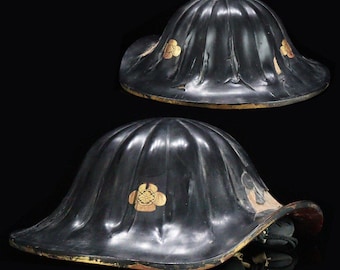May include: Two black lacquered Japanese samurai helmets, known as jingasa, featuring a fluted, ribbed dome shape and a wide, curved brim. Each helmet is decorated with a gold-colored floral kamon emblem on the side and has a red interior lining.