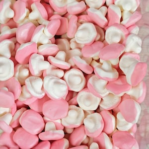 May include: A large collection of mushroom-shaped gummy sweets featuring a soft pink cap and a white stem. Each individual sweet has a smooth, rounded top and a distinct stalk, creating a dense pile of small, sweet, chewy confectionery treats.