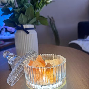 May include: A clear, ribbed glass Himalayan salt lamp with orange salt crystals. The lamp sits on a white base and is next to a white vase with white roses. The lamp's lid is off to the side.