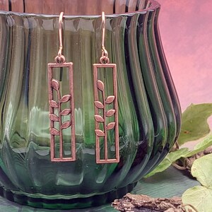 May include: Copper-coloured drop earrings. Each earring has a rectangular frame with a delicate leaf design. The earrings are displayed in front of a green glass vase against a pink and purple background.