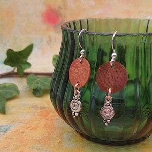 May include: Handmade earrings with a rustic design. Each earring has a circular, textured, copper-coloured pendant hanging from a silver-toned hook. Below the pendant is a decorative silver-toned charm. The earrings are displayed in a green glass vase.