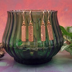 May include: Four pairs of copper-coloured rectangular dangle earrings with textured surfaces hang in front of a green glass vase. The earrings have a simple, modern design. The background is a gradient of pink and purple.