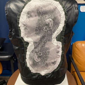 May include: Black leather jacket with a unique, artistic design on the back. The design features a grayscale image of a woman's profile, with intricate details and a white border. The jacket is displayed on a stand.