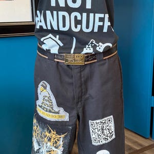 May include: A black t-shirt with the white text HOUSING NOT HANDCUFFS and a house icon, paired with black pants featuring a snake patch, a QR code, a skeleton illustration, and a large graphic print. A red beaded necklace and a belt are included.
