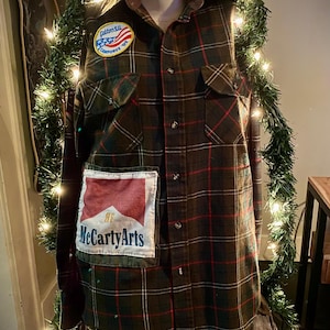 May include: A brown, green, and red plaid flannel shirt with two front pockets and a collar. Patches include a circular patch with text and an American flag design, and a rectangular patch with the text "McCartyArts".