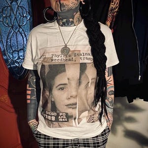 May include: White t-shirt featuring a vintage-style graphic of a woman's face with text that reads "Phyllis Stalnaker, weedhead, tramp." The shirt also includes "Diego PD APR. 1944." The person is wearing black and white plaid pants.