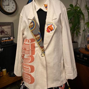 May include: A cream-colored blazer with a black shirt underneath, featuring a "Mayor McCheese" patch and a colorful sandwich-themed design. The blazer has the word "JUICE" printed in orange on the front.