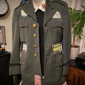 May include: An olive green military-style jacket with gold buttons and patches. The jacket has multiple pockets and is displayed on a mannequin. Patches include the text "REMEMBER" and "ONE".