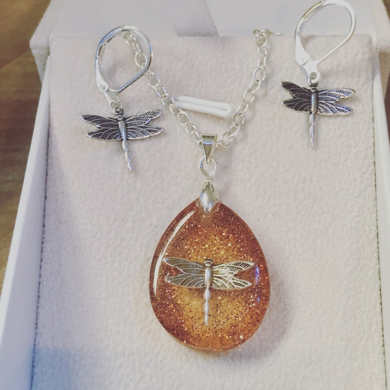 May include: A silver necklace and earrings set with a teardrop-shaped pendant featuring a glittery orange resin with a silver dragonfly inside. The earrings are also silver with dragonfly charms.