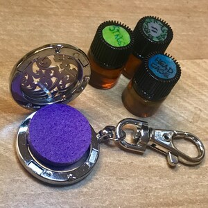 Könnte beinhalten: Ein silbernes Medaillon mit einem violetten Filzpad im Inneren, geöffnet, um das Pad zu zeigen. Das Medaillon ist an einem silbernen Schlüsselanhänger befestigt. Im Hintergrund befinden sich drei kleine Glasflaschen mit schwarzen Kappen. Die Flaschen sind mit "Sting", "Head Space" und "Sage" beschriftet.