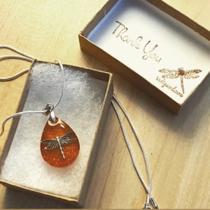 May include: A silver chain necklace with an orange teardrop pendant featuring a silver dragonfly. The necklace is in a small brown box with white tissue paper. The box has a thank you message and a dragonfly logo.
