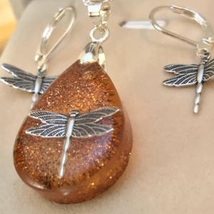 May include: A teardrop-shaped pendant and matching earrings feature a silver dragonfly design set in clear resin with gold glitter.