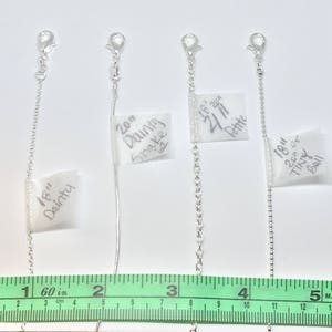 May include: Four silver chains of different lengths and styles, labeled with their measurements and descriptions. The chains are laid out on a white surface with a green ruler underneath. The labels read: 18" Dainty, 20" Dainty Snake, 18" 20" Petite, 18" 20" Tiny Ball.