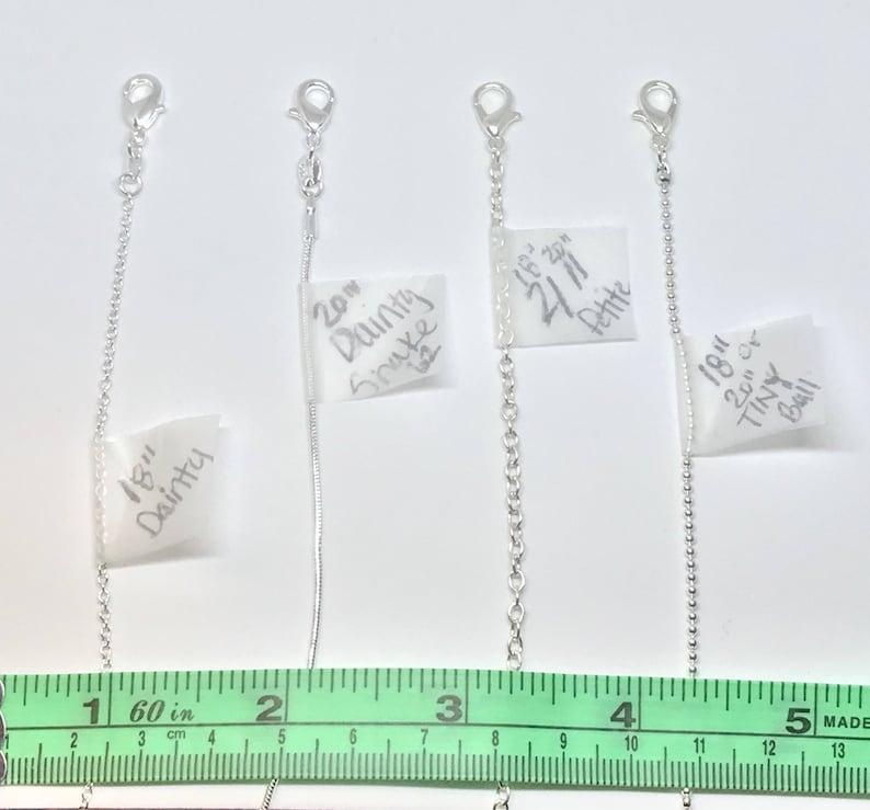 May include: Four silver chains of different lengths and styles, labeled with their measurements and descriptions. The chains are laid out on a white surface with a green ruler underneath. The labels read: 18" Dainty, 20" Dainty Snake, 18" 20" Petite, 18" 20" Tiny Ball.