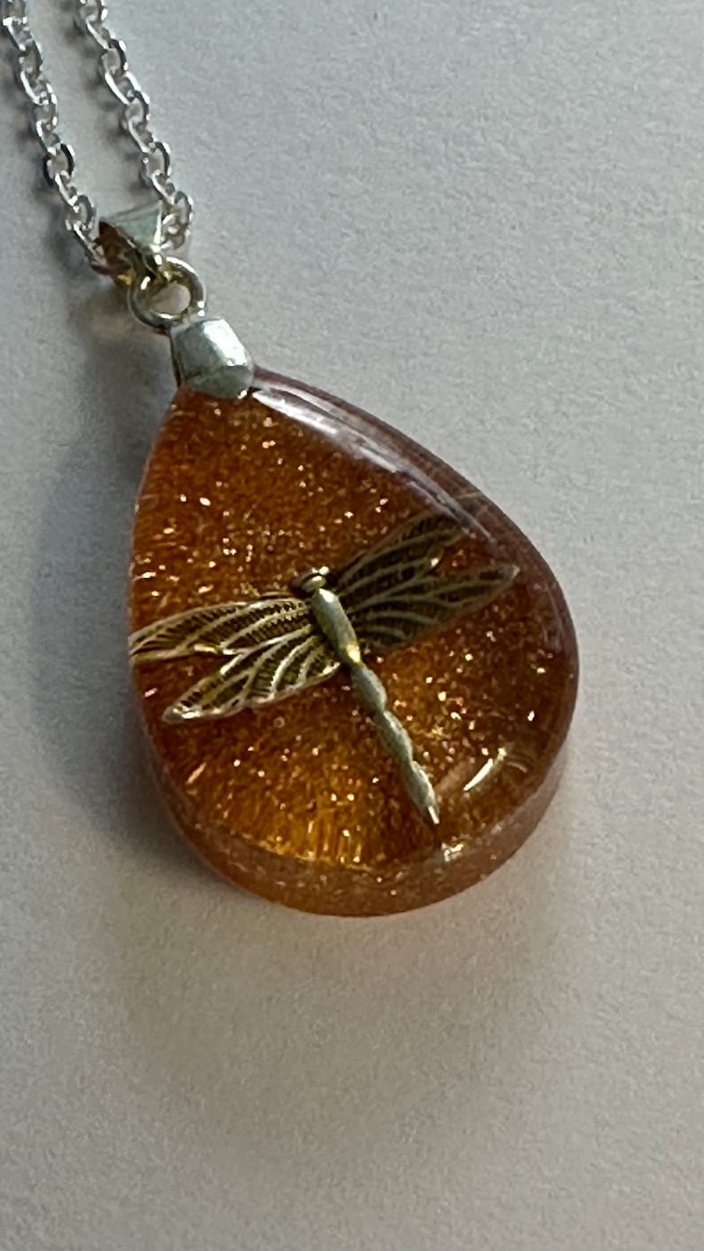 May include: A gold dragonfly pendant necklace with a teardrop-shaped resin charm filled with gold glitter.