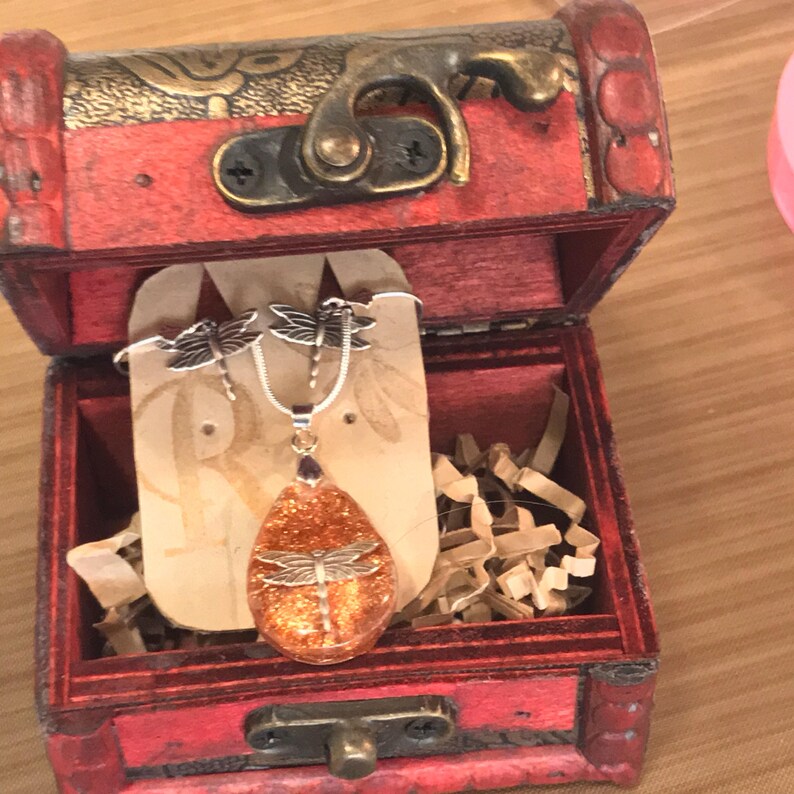 May include: A set of dragonfly jewelry in a red wooden box. The necklace features a teardrop-shaped pendant with a dragonfly encased in orange glitter resin. The earrings are silver with dragonfly accents.