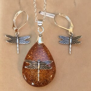 May include: A silver necklace and earrings set with a teardrop-shaped pendant filled with gold glitter and a dragonfly charm. The dragonfly charm is also on the earrings.