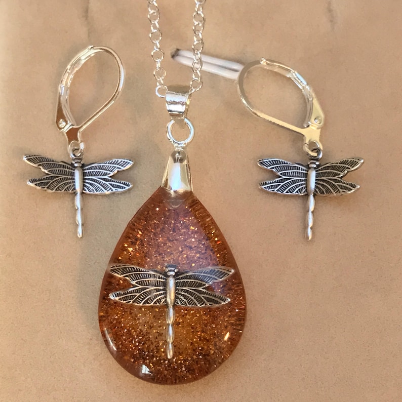 May include: A silver necklace and earrings set with a teardrop-shaped pendant filled with gold glitter and a dragonfly charm. The dragonfly charm is also on the earrings.