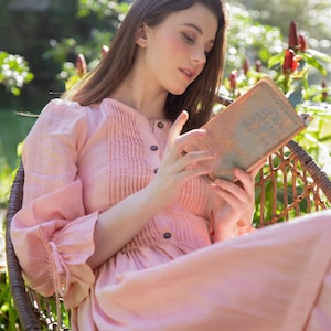 Könnte beinhalten: Eine Frau in einem rosa Kleid sitzt im Freien auf einem Korbstuhl und liest ein Buch. Das Kleid hat einen Knopfleisten vorne und eine geraffte Taille.